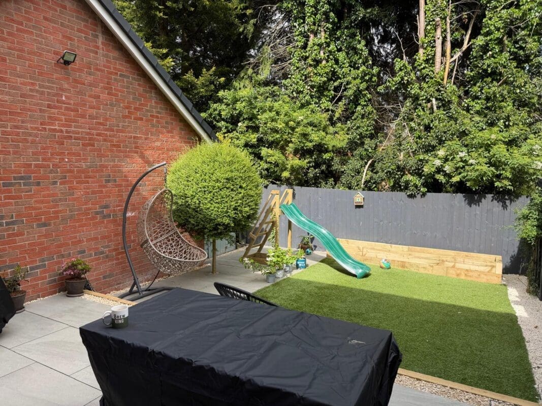 Family garden in Worcestershire showing artificial lawn, timber sandpit, green children’s slide, and hanging egg chair against dark grey fence panels